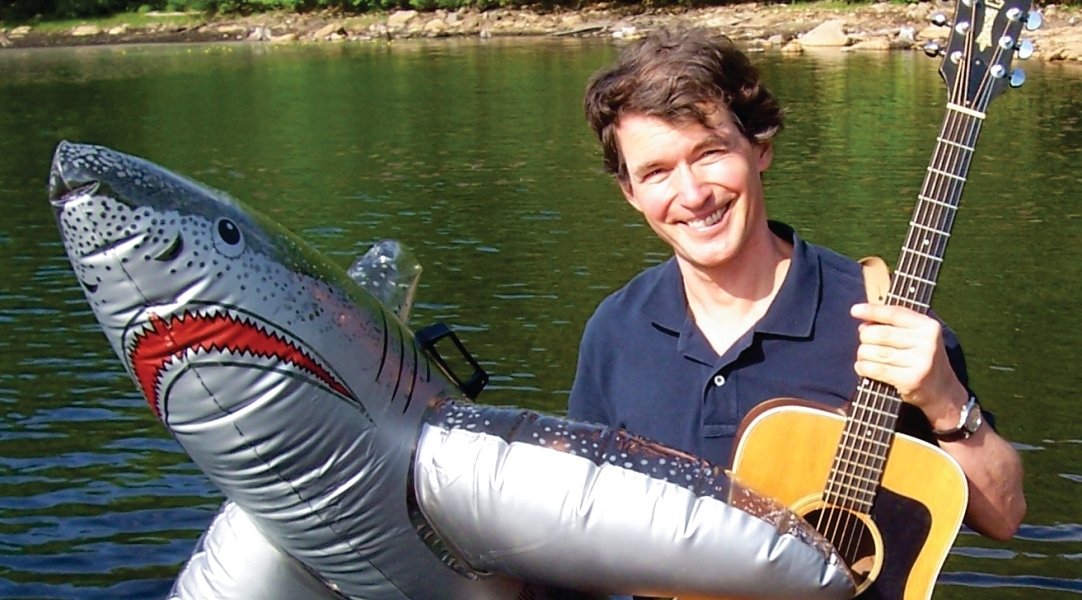 Tom Seiling with his guitar and an inflatable shark standing in front of a pond