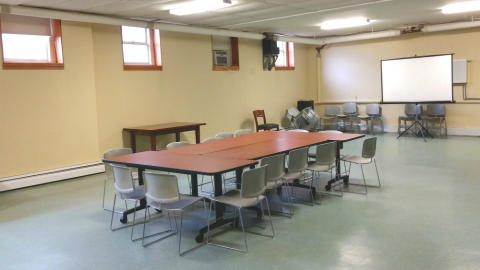 Photograph of the interior of the Forest Park branch library community room with tables, chairs, and projector screen. Small windows.