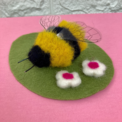 fuzzy felted black and yellow bee sitting on a green felt leaf with two small felted white and pink flowers