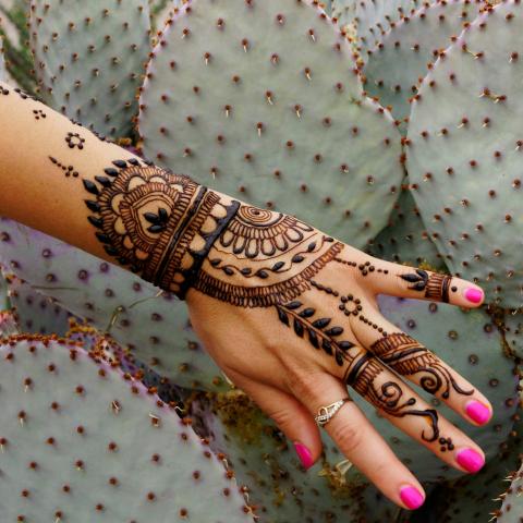 A hand with henna art and pink fingernails in front of a backdrop of cactuses.