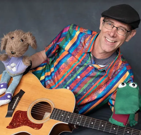 Tom Knight in a colorful shirt with two puppets and a guitar.