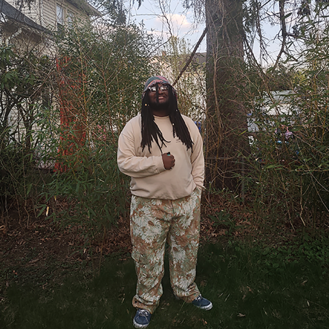 The performer Specifix, a black man with long locs, standing in a wooded area