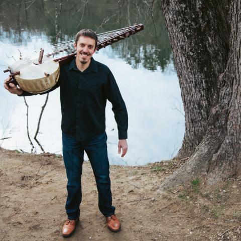 A young man holding a long stringed instrument, the kora, in front of a bost of water and a large tree trunk. 