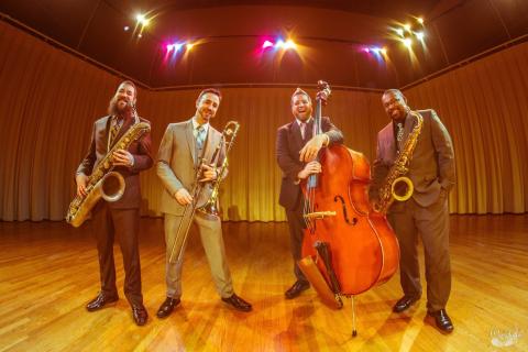 Flava Evolution, a 4-man band with bari and alto sax, trombone, and upright bass. They're standing on a stage with multicolored lights and there is a slight fisheye effect