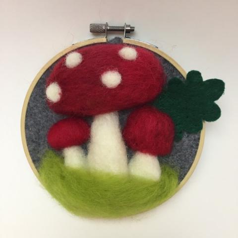 Red and white felted toadstools on green felt grass in a small embroidery hoop