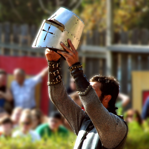 An armored knight hoists his helmet into the hair before a crowd of onlookers.