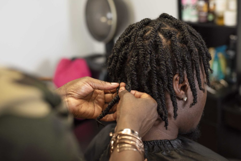 The back of a person's head. Their Black hair is being braided by someone wearing gold bracelets.