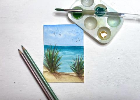 small water color painting of beach scene, with pencils and watercolor paints nearby on the table