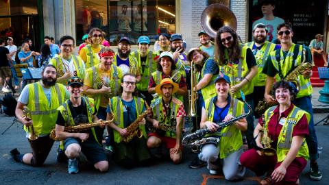 A large group of band members with brass instruments. They're all wearing yellow hi-vis safety vests. Many of them have cultural references in their attire to shows like One Piece and MST3K.