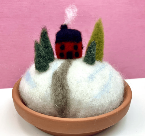 Felted 3-d scene in a small flowerpot of snowy field with a tall red barn and green fir trees along a grey road