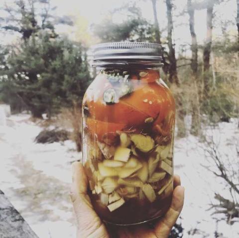 A hand holding a large glass jar of ambe liquid with chunks of vegetation, in front of a snowy outdoor woodland background