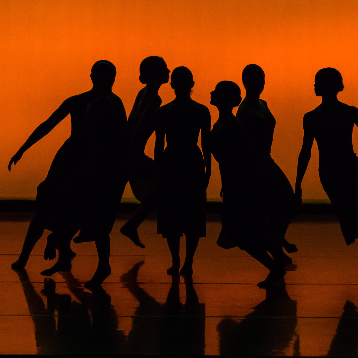 Silhouettes of six figures dancing on a stage with a vibrant orange background.