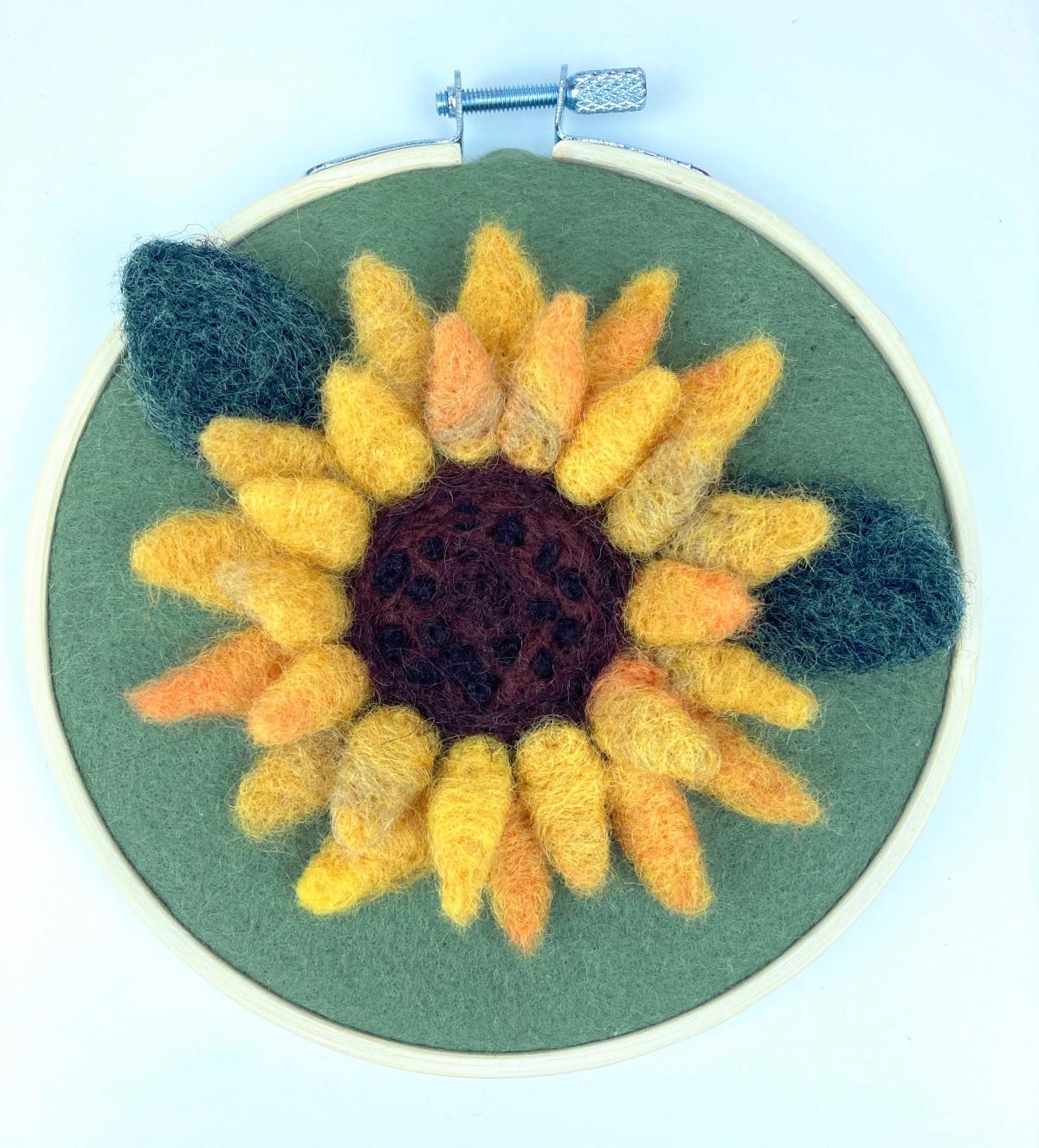 Felted sunflower in shades of yellow with a black center on a green felt background in a hoop