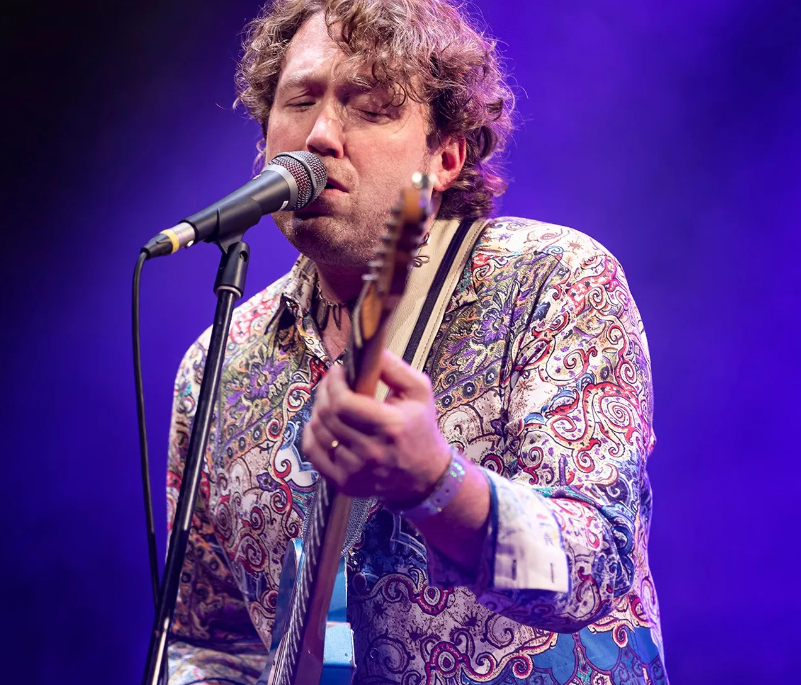 A man singing into a microphone and holding an electric guitar, on a blue lit background